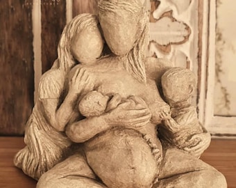 Op de afbeelding: Een beige sculptuur die een zwangere persoon voorstelt die zit met twee kinderen en een pasgeborene. De sculptuur heeft een getextureerde afwerking en staat tegen een houten achtergrond. De tekst "moon birth" is zichtbaar.