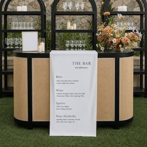 Könnte beinhalten: Eine Bar mit einem weißen Menütuch mit der Aufschrift "THE BAR menu". Die Bar hat eine Vorderseite aus gewebtem Rattan und schwarze Akzente. Regale beherbergen Gläser, Kerzen und Blumengestecke. Die Speisekarte listet Bier, Wein, Spirituosen und alkoholfreie Getränke auf.