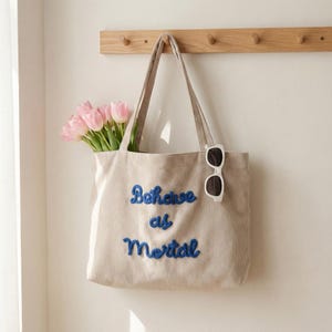 Peut inclure: Sac fourre-tout en velours côtelé beige avec le texte brodé en bleu "Behave as Mortal". Le sac est suspendu à un porte-manteau en bois et contient un bouquet de tulipes roses et une paire de lunettes de soleil blanches.