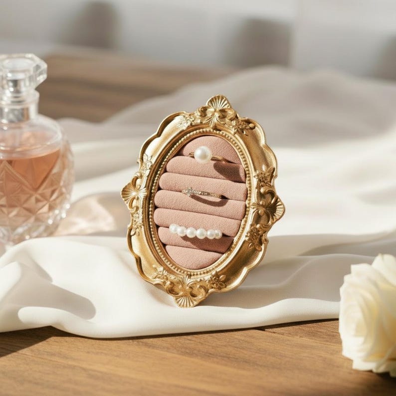 May include: A gold-colored, ornate oval ring holder with a pink velvet interior, displaying three rings. One ring features a pearl, another has small stones, and the third has multiple pearls. A perfume bottle is in the background.