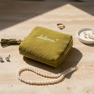 May include: An olive green jewellery case embroidered with the name "Selena" in white, a gold zip, and a tassel. A pearl necklace, a gold ring, and a small dish with pearls are also visible.