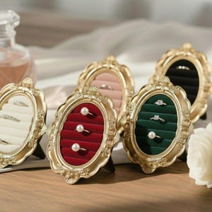 May include: Ornate gold-colored ring displays with oval openings. Each display features a velvet interior in various colors: white, pink, red, green, and black. Some displays hold rings with pearls or gemstones. The displays are set on a wooden surface.