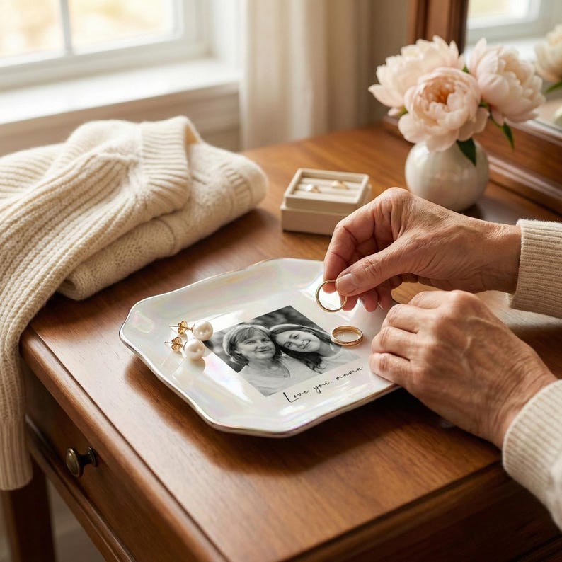 May include: A white, iridescent jewelry tray with a gold trim, holding a black and white photo and two gold rings. The tray also holds a pair of pearl earrings. A folded cream sweater and a small vase of pink flowers are in the background.