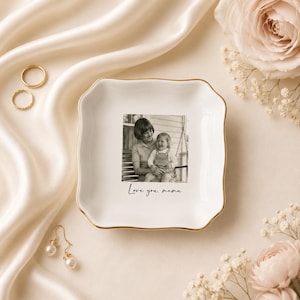 May include: A white ceramic trinket dish with a gold rim and scalloped edges. The dish features a black and white photograph of a mother and child, with the text "Love you, mama." Gold rings and pearl earrings are also visible.