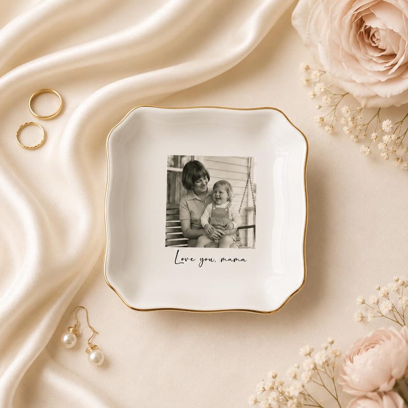 May include: A white ceramic trinket dish with a gold rim and scalloped edges. The dish features a black and white photograph of a mother and child, with the text "Love you, mama." Gold rings and pearl earrings are also visible.