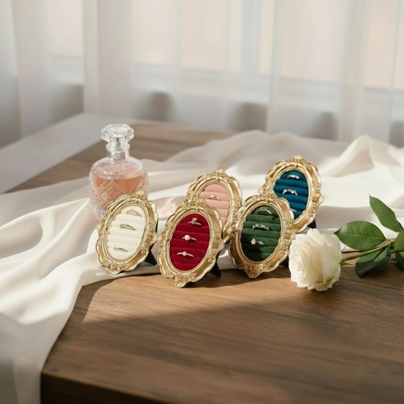 May include: Oval-shaped ring display cases with gold frames and velvet interiors in cream, pink, red, green, and teal. A perfume bottle and a white rose are also in the image. The cases are on a wooden surface.
