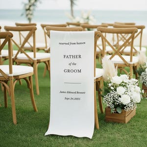 May include: A white fabric sign draped over a wooden cross-back chair, featuring black text that reads: reserved from heaven, FATHER of the GROOM, James Edward Bennett, Sept. 24, 2025. The chair is placed on green grass near a wooden box of white flowers.