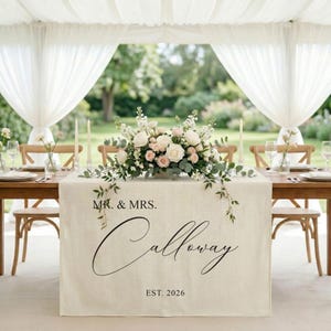 May include: A beige linen table runner draped over a table, featuring the black printed text MR. & MRS. Calloway EST. 2026. A large floral centrepiece with white and light pink roses and green eucalyptus leaves sits on top of the table runner.