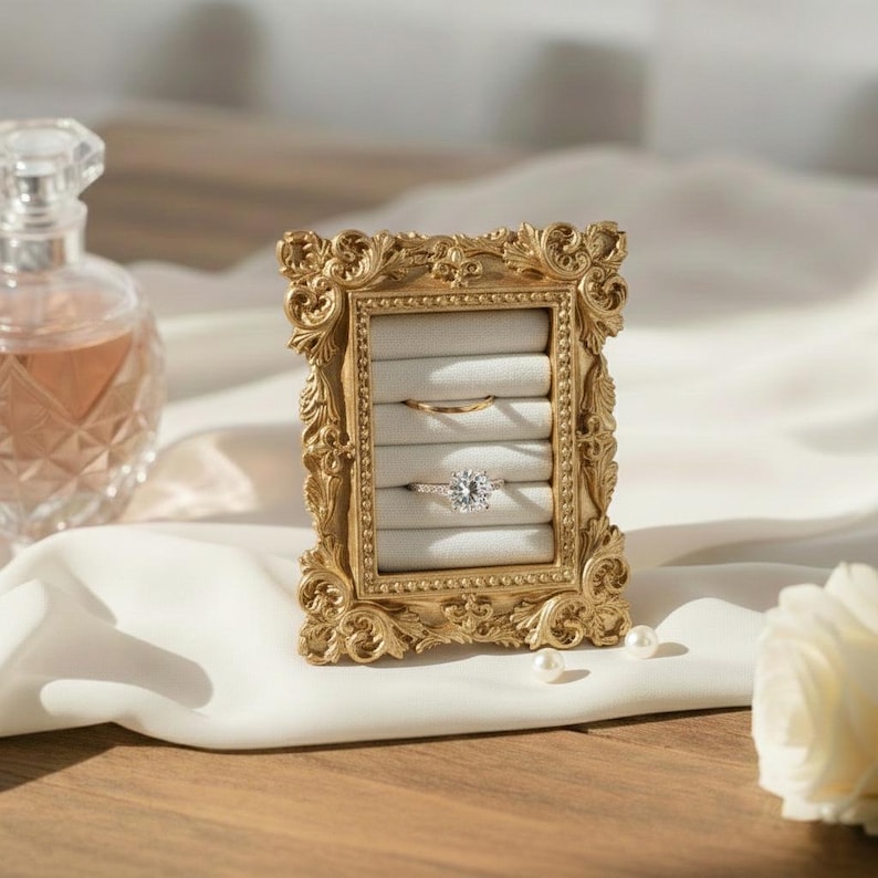 May include: A gold-colored, ornate picture frame holds two rings on a gray velvet insert. The frame has an intricate, detailed design. A diamond ring and a gold band are displayed. The background includes a perfume bottle and white fabric.