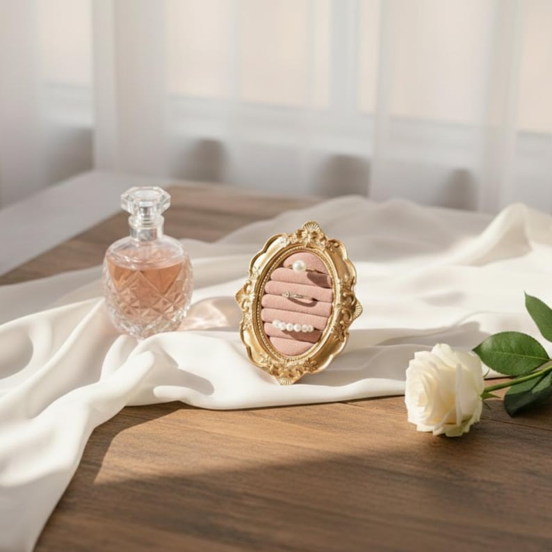 May include: A gold-colored, ornate oval jewelry display with a pink velvet interior holds rings and pearls. A clear perfume bottle and a white rose rest on a wooden surface with a white fabric backdrop.