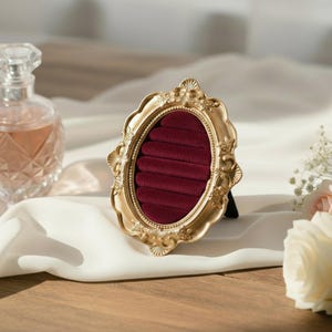 May include: Ornate gold-colored jewelry display with a burgundy velvet interior. The oval-shaped frame features intricate detailing and a small black stand. A perfume bottle and flowers are in the background.