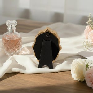 May include: A gold-colored, ornate picture frame with a black backing and a black easel stand. A clear perfume bottle with a diamond pattern and pink liquid is to the left. Soft pink and white roses are in the background.