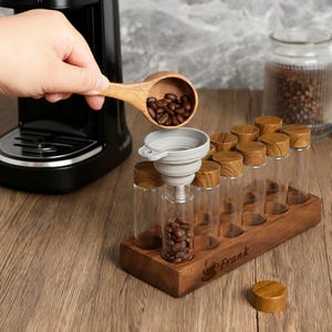 May include: A wooden scoop pours coffee beans into a gray funnel, filling a clear glass vial. The vial is part of a set of ten vials with wooden lids, displayed in a wooden holder. A coffee maker is in the background.