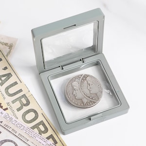Puede incluir: Una moneda de plata con dos perfiles, exhibida en una vitrina gris con bisagras. La moneda presenta el año 1921 y la inscripción "E PLURIBUS UNUM". La vitrina está abierta, revelando la moneda sobre un fondo blanco.
