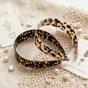Puede incluir: Dos diademas con estampado de carey. Una es marrón oscuro y la otra beige claro con manchas marrones oscuras. Las diademas están sobre una superficie color crema con pequeñas perlas y una tarjeta que dice "Summer at the Coast".