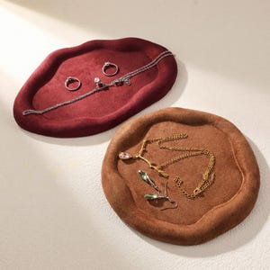 May include: Two jewelry trays in the image. One is a deep red, oval-shaped tray with rings and a necklace. The other is a round, brown tray with necklaces and earrings. The trays are made of a soft, velvety material.