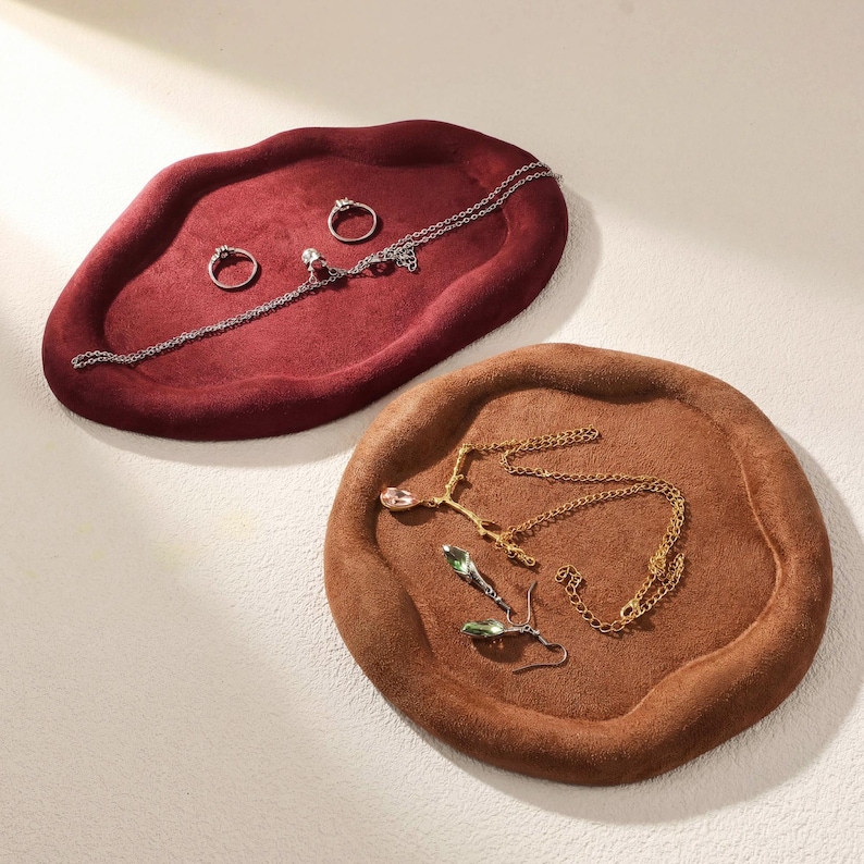 May include: Two jewelry trays in the image. One is a deep red, oval-shaped tray with rings and a necklace. The other is a round, brown tray with necklaces and earrings. The trays are made of a soft, velvety material.