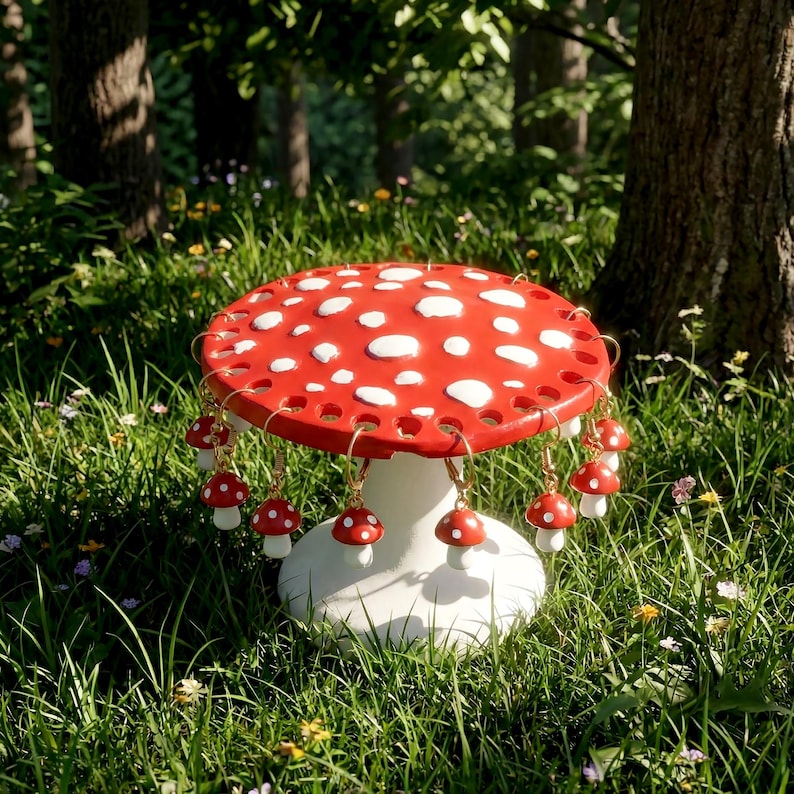 May include: A whimsical mushroom-shaped table with a red top and white spots, adorned with dangling mushroom earrings. The table is white and sits in a grassy area with trees in the background. The earrings have gold-colored hooks.