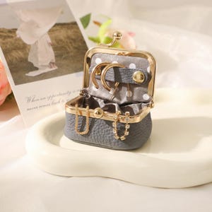 Puede incluir: Una pequeña caja de joyería gris en forma de monedero, con un marco y cierre de color dorado. El interior está forrado con tela gris y contiene aretes y collares dorados. Una tarjeta decorativa está en el fondo.