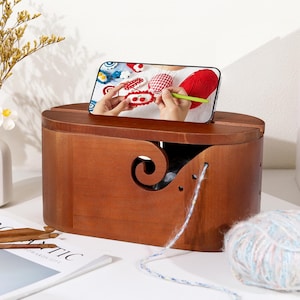 May include: A wooden yarn bowl with a phone displaying a crochet tutorial. The bowl is a warm brown color with a curved design and a swirl cutout. A ball of yarn and crochet hooks are nearby, suggesting a crafting theme.