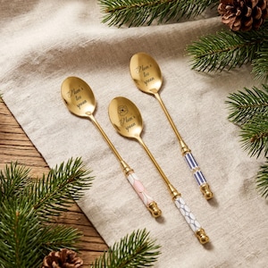 Può includere: Tre cucchiaini da tè dorati con la scritta "Mom's tea spoon" incisa sulle teste dei cucchiai. Due cucchiai hanno manici fantasia rosa e bianchi, e blu e bianchi. I cucchiai sono disposti su un panno di colore neutro.