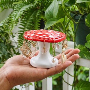 May include: A red and white mushroom-shaped earring holder. The top is red with white spots, and the base is white. Various earrings, including gold and crystal designs, hang from the edges. The holder is held in a hand.