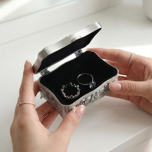 Puede incluir: Una pequeña caja de joyería plateada con la tapa abierta, que revela un interior negro. En el interior, se ven dos anillos: uno con un diseño circular y otro con una piedra oscura. La caja es sostenida por manos.