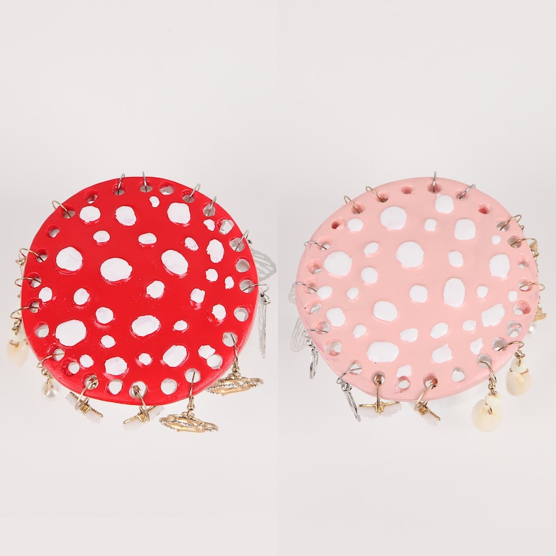 May include: Two mushroom-shaped earrings. One is red with white spots, the other is pink with white spots. Both have dangling charms and are designed to be worn as earrings. The earrings are a whimsical accessory.