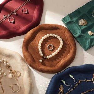 May include: An overhead shot of jewelry displayed on velvet trays in various colors. A pearl bracelet and ring sit on a brown tray. Other trays are burgundy, cream, green, and navy blue, each holding different jewelry pieces like necklaces and earrings.