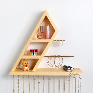 Peut inclure: Un organisateur de bijoux mural en bois de forme géométrique. Il comprend des étagères avec des flacons de parfum, un petit pot et une figurine d'ours en peluche. En dessous, des barres métalliques pour boucles d'oreilles et bracelets, ainsi que des crochets pour colliers. Le nom "Sophia" est gravé en bas.