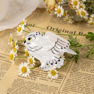 May include: A white owl figurine with black detailing and yellow eyes rests among daisies on a vintage newspaper. The owl has a pearlescent finish, and its wings are spread. The overall theme is nature and decor.