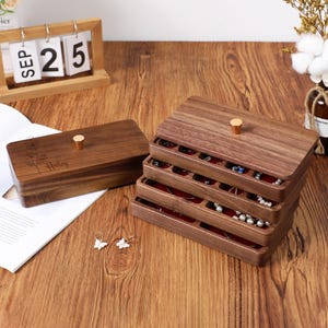 Peut inclure: Coffret à bijoux en bois foncé avec plusieurs tiroirs doublés de velours rouge. Le coffret a un bouton doré sur le couvercle. Une petite boîte avec le nom "Haley" gravé est également présente. Le coffret à bijoux est sur une surface en bois.