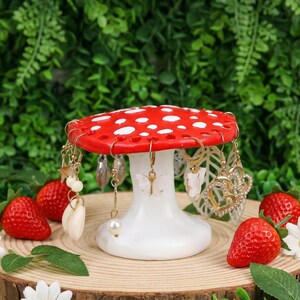 May include: A whimsical red and white mushroom-shaped earring holder. The holder is adorned with various earrings, including gold-toned hoops, pearl drops, and butterfly designs. Strawberries and greenery surround the holder, creating a charming display.