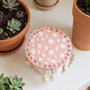 May include: A pink ceramic jewelry holder with white polka dots. The holder has small holes around the edge for hanging earrings. Several pairs of earrings are displayed. The holder sits on a white surface, with potted plants in the background.