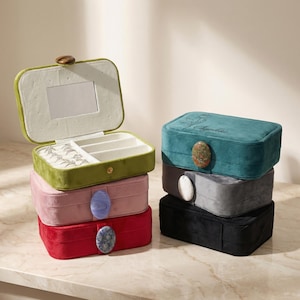 May include: A stack of velvet jewelry boxes in various colors: olive green, pink, red, gray, teal, and black. Each box has a decorative stone clasp. The top box is open, revealing a mirror and compartments for jewelry.