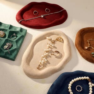 May include: An assortment of jewelry displayed on velvet trays in various colors. The trays hold rings, bracelets, and necklaces. The trays are in shades of red, green, beige, brown, and blue. The jewelry includes pearls, gold, and silver pieces.