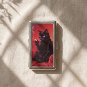 Può includere: Un portasigarette rettangolare in metallo argentato con l'immagine stampata di un gatto nero seduto su una superficie rossa, che tiene in mano una sigaretta accesa con fumo visibile. L'astuccio presenta una cornice metallica e una chiusura laterale.