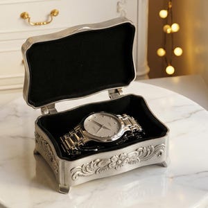 May include: An open, ornate silver jewelry box with a black velvet interior holds a silver wristwatch. The box features floral and scrollwork designs. The watch has a silver band and a light-colored face.