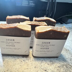 Op de afbeelding: Vier rechthoekige zeepblokken met een donkerbruine basis en een lichtere bruine bovenkant. Elk blok is verpakt met een wit etiket met de tekst "SOVON HONEY, MOLASSES, COCONUT GLOW LUXE SOAP BAR". De zeepblokken wegen ongeveer 127g per stuk.