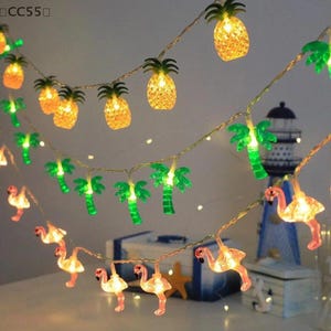 May include: String lights with pineapple, palm tree, and flamingo-shaped lights. The pineapple lights are yellow, the palm trees are green, and the flamingos are pink. The lights are strung on a thin, brown wire.
