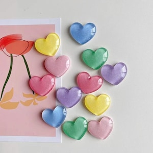 Puede incluir: Una colección de adornos en forma de corazón de varios colores. Los corazones son en tonos rosa, amarillo, azul, verde y morado. Parecen translúcidos con un ligero brillo. Los corazones están dispuestos sobre una superficie blanca y rosa.