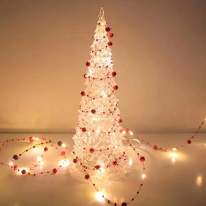 May include: A clear, illuminated Christmas tree with red bead garland. The tree is lit from within, creating a warm glow. A string of lights with red beads extends from the tree.