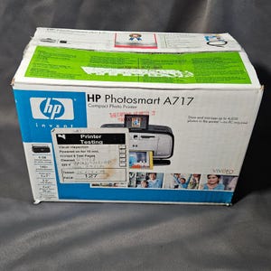 May include: An HP Photosmart A717 compact photo printer in its original box. The box is white with blue accents and features images of the printer and photos. Text on the box includes "Printer Testing" and "Store and manage up to 4,000 photos."