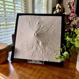 May include: A framed, textured artwork depicting Mount Fuji. The artwork is in a black frame and features the mountain's form in a light, neutral color. The text "Mount Fuji" and coordinates are visible at the bottom.
