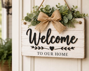 Può includere: Un cartello rettangolare in legno bianco con finitura anticata, che presenta la scritta nera in 3D Welcome e il testo TO OUR HOME. È decorato con un fiocco in juta e foglie di eucalipto artificiali, appeso a una corda marrone.