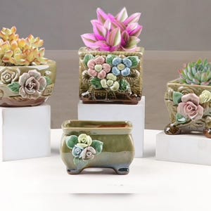 May include: A collection of ceramic flower pots in various shapes, each adorned with embossed floral designs and filled with succulents. The pots are in shades of green and brown, with pink, blue, and yellow flowers. They sit on white pedestals.
