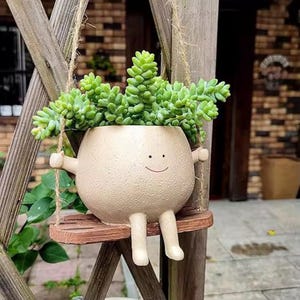May include: A beige ceramic planter shaped like a smiling character, with arms and legs, suspended on a wooden swing. The planter is filled with green succulent plants. The swing is hanging from a wooden structure.