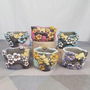 May include: Six small, decorative planters with a bear and floral design. Each planter is a different color, including pink, blue, and gray, and features a tree branch with flowers and a small bear. The planters are made of a textured material.