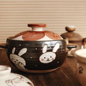 Può includere: Una pentola in ceramica rotonda con un esterno marrone scuro a coste, decorata con illustrazioni di conigli bianchi e piccoli punti bianchi. È dotata di due manici laterali e di un coperchio abbinato con un motivo circolare marrone e bianco.