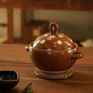 Peut inclure: Un bol en céramique marron avec couvercle, doté d'une finition brillante et d'un motif à pois blancs sur le couvercle. Le bol possède deux anses latérales et une poignée en forme de boucle sur le dessus. Il repose sur un dessous de plat tressé.